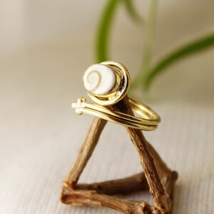 K&ouml;nnte beinhalten: Goldring mit einer wei&szlig;en Muschel mit einem Spiralmuster. Der Ring wird auf einem Holzst&auml;nder pr&auml;sentiert.