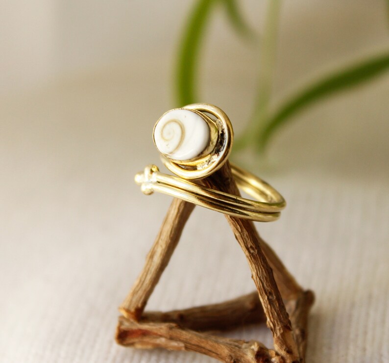 K&ouml;nnte beinhalten: Goldring mit einer wei&szlig;en Muschel mit einem Spiralmuster. Der Ring wird auf einem Holzst&auml;nder pr&auml;sentiert.