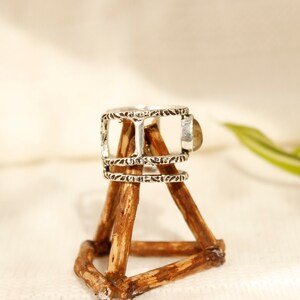 May include: Silver ring with a decorative design and a light brown stone. The ring is displayed on a wooden stand.