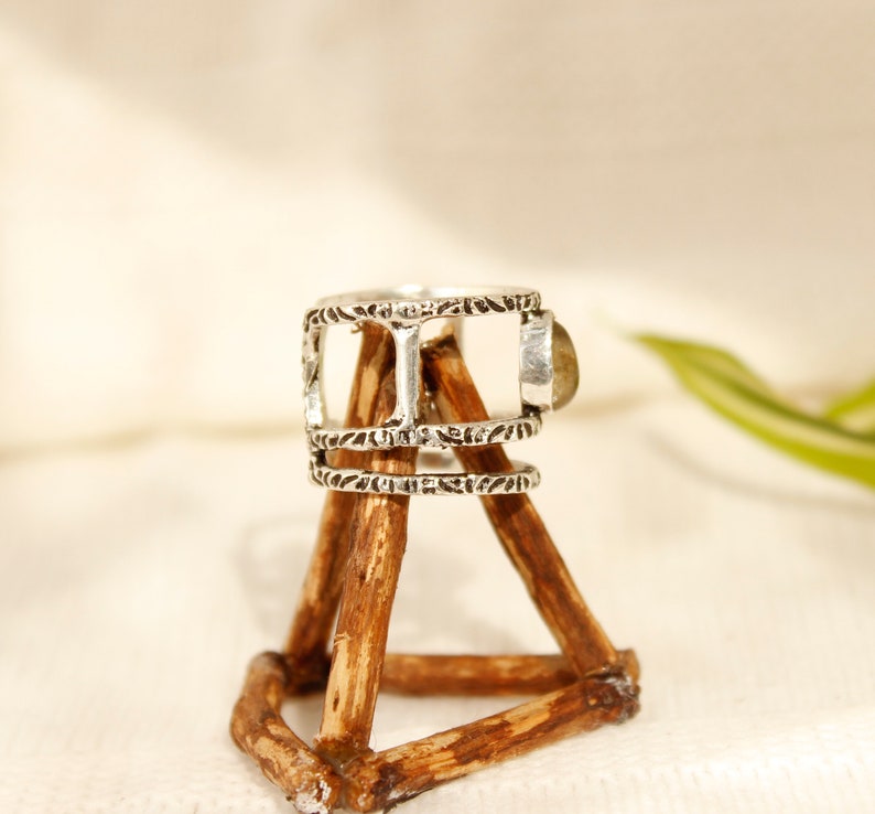 May include: Silver ring with a decorative design and a light brown stone. The ring is displayed on a wooden stand.