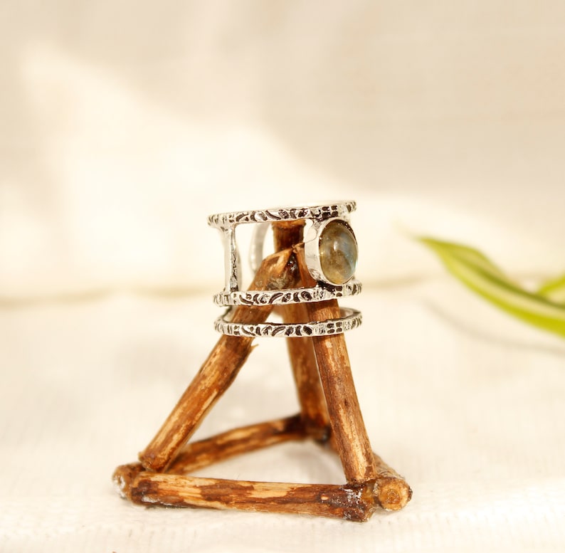 May include: Silver ring with a labradorite gemstone set in a triple band design. The ring is displayed on a wooden stand.