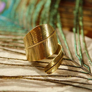 Könnte beinhalten: Ein goldfarbener Ring in Form einer Feder. Der Ring umschlingt den Finger mit einem detaillierten Federdesign. Der Ring ist vor dem Hintergrund einer Pfauenfeder platziert.