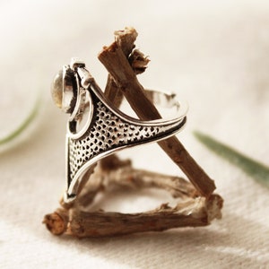 May include: A silver ring with a delicate, intricate design and a clear gemstone. The ring is resting on a piece of driftwood.