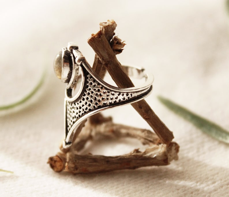 May include: A silver ring with a delicate, intricate design and a clear gemstone. The ring is resting on a piece of driftwood.
