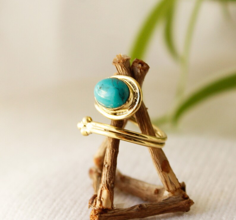 K&ouml;nnte beinhalten: Ein goldener Ring mit einem t&uuml;rkisfarbenen Stein, der in einem gedrehten Band gefasst ist. Der Ring wird auf einem Holzst&auml;nder pr&auml;sentiert.