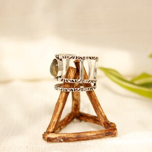 May include: Silver ring with a green gemstone set in a decorative band. The ring is displayed on a wooden stand.
