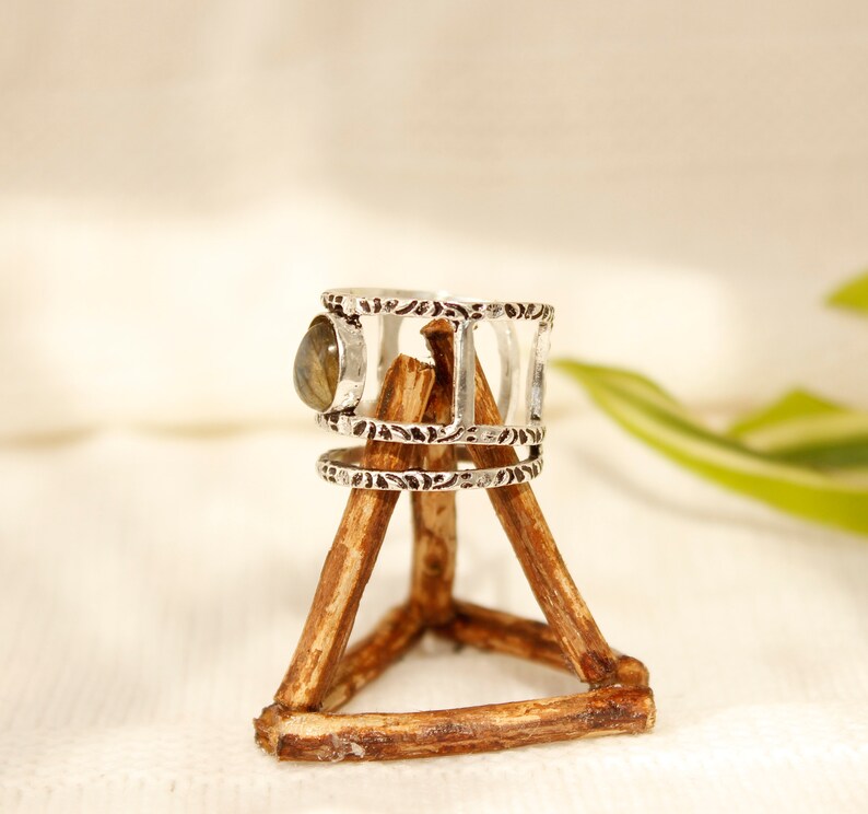 May include: Silver ring with a green gemstone set in a decorative band. The ring is displayed on a wooden stand.
