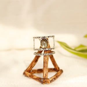 May include: A silver ring with a light gray oval gemstone set in a textured band. The ring is displayed on a wooden ring holder.