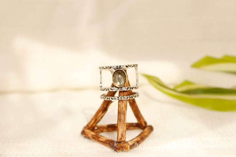 May include: A silver ring with a light gray oval gemstone set in a textured band. The ring is displayed on a wooden ring holder.
