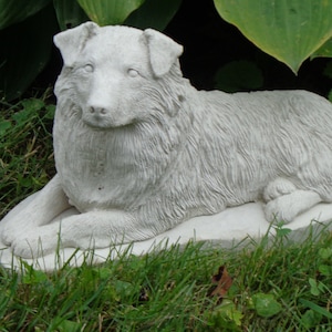 May include: A white concrete statue of a dog lying down. The dog is sculpted in a realistic style with fur details. The statue is placed on a grassy surface.