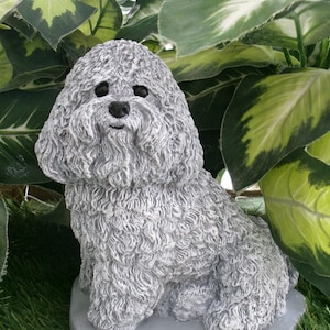 May include: A gray concrete statue of a sitting Bichon Frise dog. The statue is on a gray base and is surrounded by green grass.