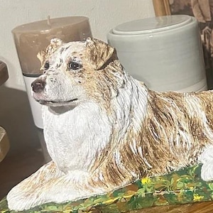 May include: A brown and white ceramic figurine of a dog lying down on a green and yellow base. The dog has a white chest and a brown and white coat.