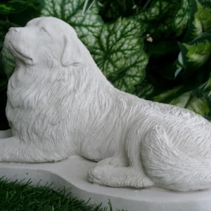 May include: A white, detailed sculpture of a dog in a relaxed pose, with its head tilted upwards. The dog is lying on a rectangular base, set against a backdrop of green foliage and grass. The sculpture has a textured surface.