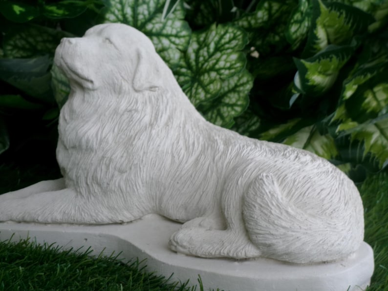 May include: A white, detailed sculpture of a dog in a relaxed pose, with its head tilted upwards. The dog is lying on a rectangular base, set against a backdrop of green foliage and grass. The sculpture has a textured surface.