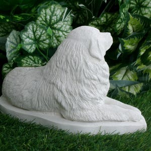 May include: A white, stone-like sculpture of a dog in a resting pose. The dog is lying down on a flat, oval base, with its head turned to the side. The sculpture has detailed fur texture and is set against a backdrop of green foliage and grass.