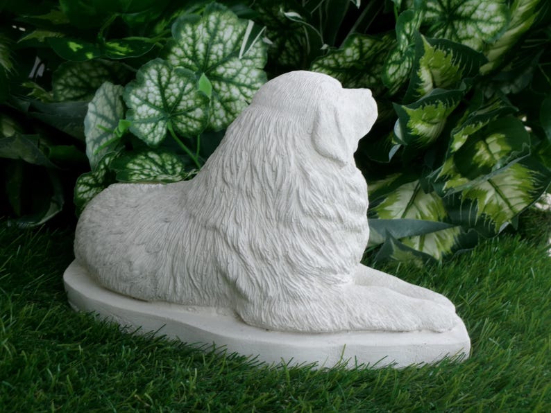 May include: A white, stone-like sculpture of a dog in a resting pose. The dog is lying down on a flat, oval base, with its head turned to the side. The sculpture has detailed fur texture and is set against a backdrop of green foliage and grass.