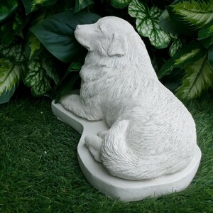 May include: A light gray dog statue in a resting pose. The dog is lying down on a matching base, with its head turned to the side. The statue is surrounded by green foliage and grass.