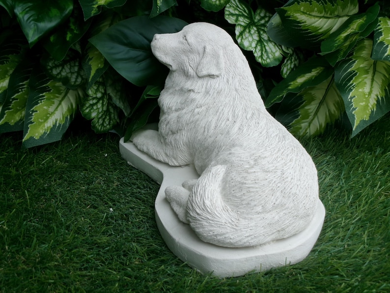 May include: A light gray dog statue in a resting pose. The dog is lying down on a matching base, with its head turned to the side. The statue is surrounded by green foliage and grass.