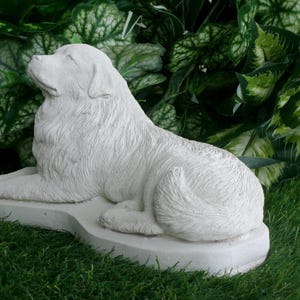 May include: A white, detailed sculpture of a dog in a resting pose. The dog is lying down on a rectangular base, with its head raised and eyes closed. The sculpture is set against a backdrop of green foliage and grass.