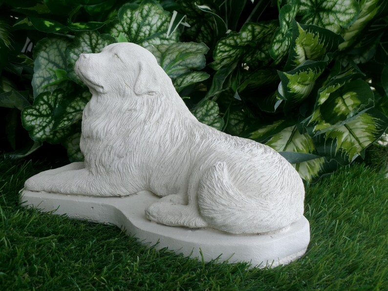 May include: A white, detailed sculpture of a dog in a resting pose. The dog is lying down on a rectangular base, with its head raised and eyes closed. The sculpture is set against a backdrop of green foliage and grass.