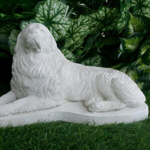 May include: A white, reclining dog statue with detailed fur texture. The sculpture is resting on a rectangular base, set against a backdrop of green foliage and grass. The dog's head is raised, and its eyes are closed.
