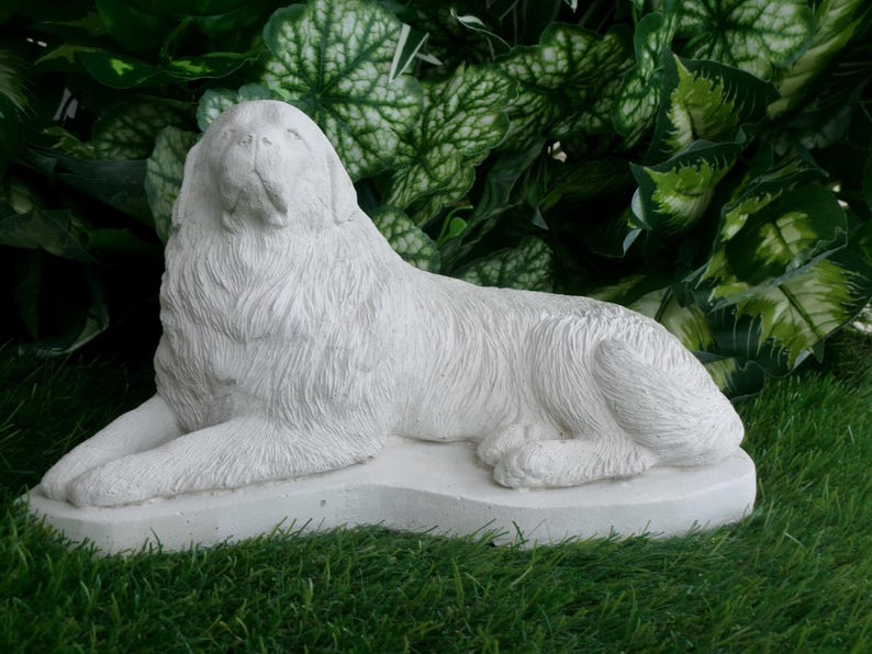 May include: A white, reclining dog statue with detailed fur texture. The sculpture is resting on a rectangular base, set against a backdrop of green foliage and grass. The dog's head is raised, and its eyes are closed.
