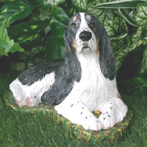 May include: A hand-painted figurine of a tri-color English Setter dog lying down. The dog features black, white, and tan fur markings, long ears, and a textured coat. It is positioned on a flat, green, and brown mottled base with a realistic finish.