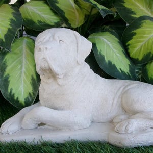 May include: A white concrete statue of a dog lying down. The dog is in a relaxed pose with its head resting on its paws. The statue is on a green grassy surface.