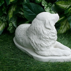 May include: A light-colored, stone-like sculpture of a dog in a resting pose. The dog is lying down with its head raised, and its fur is textured. The sculpture is on a base and set against a backdrop of green foliage.