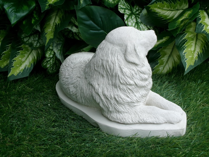 May include: A light-colored, stone-like sculpture of a dog in a resting pose. The dog is lying down with its head raised, and its fur is textured. The sculpture is on a base and set against a backdrop of green foliage.