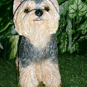 May include: A sculpted figurine of a Yorkshire Terrier dog with a textured coat in shades of tan, black, and grey. The dog is depicted in a seated position, featuring upright ears, dark eyes, and a black nose, mounted on a base.