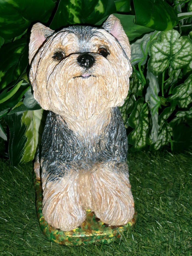 May include: A sculpted figurine of a Yorkshire Terrier dog with a textured coat in shades of tan, black, and grey. The dog is depicted in a seated position, featuring upright ears, dark eyes, and a black nose, mounted on a base.