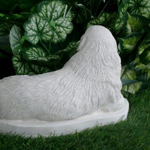 May include: A white, textured statue of a dog lying down, viewed from the back. The sculpture is set on a circular base and is surrounded by green foliage and grass.