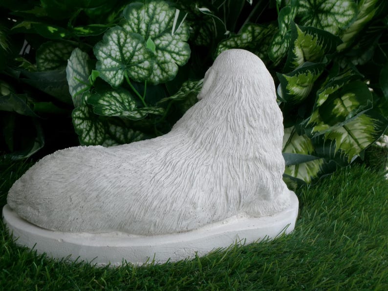 May include: A white, textured statue of a dog lying down, viewed from the back. The sculpture is set on a circular base and is surrounded by green foliage and grass.