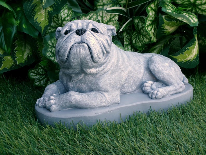 Może przedstawiać: Szara statua buldoga w zrelaksowanej pozie, spoczywająca na szarej podstawie. Rzeźba ma szczeg&oacute;łowe zmarszczki i poważny wyraz twarzy. Statua jest ustawiona na tle zielonych liści i trawy.