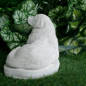 May include: A white, seated dog statue with textured fur detail. The sculpture is on a round base and set against a backdrop of green foliage and grass. The dog is facing away from the viewer.