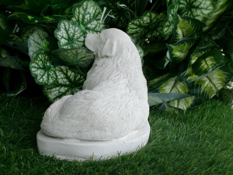 May include: A white, seated dog statue with textured fur detail. The sculpture is on a round base and set against a backdrop of green foliage and grass. The dog is facing away from the viewer.