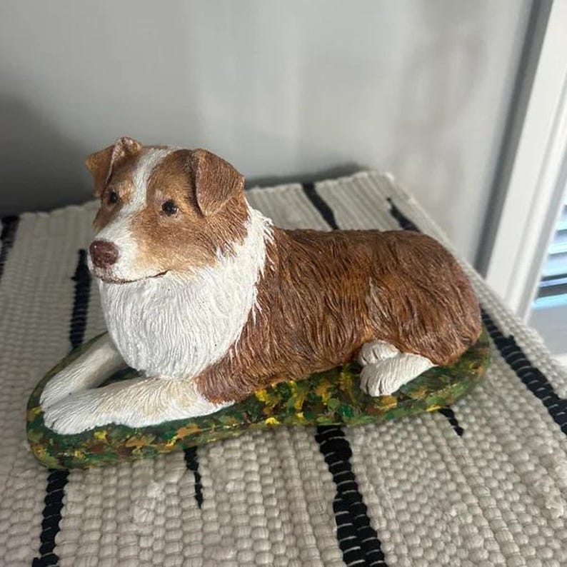 May include: A brown and white figurine of an Australian Shepherd dog lying down on a green and brown base. The dog has a white chest and a white patch on its head.