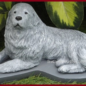 May include: A gray concrete statue of a dog, lying down with its front paws extended. The dog has a fluffy coat and is resting on a gray base. The statue is outdoors, with green foliage in the background.