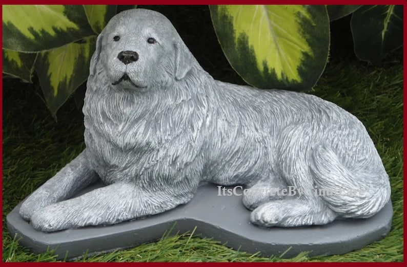 May include: A gray concrete statue of a dog, lying down with its front paws extended. The dog has a fluffy coat and is resting on a gray base. The statue is outdoors, with green foliage in the background.