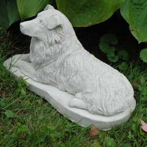 May include: A white concrete statue of a dog lying down on a base. The dog is sculpted in a realistic style with fur details.