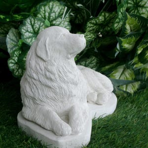 May include: A white, detailed sculpture of a dog in a relaxed pose. The dog is lying down with its head raised, and its fur is textured. The sculpture is set on a white base and surrounded by green foliage and grass.