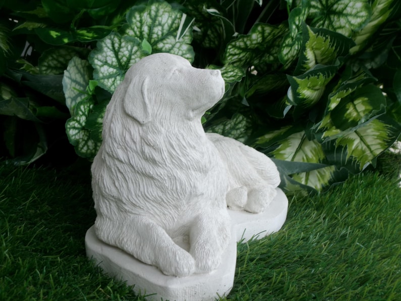 May include: A white, detailed sculpture of a dog in a relaxed pose. The dog is lying down with its head raised, and its fur is textured. The sculpture is set on a white base and surrounded by green foliage and grass.