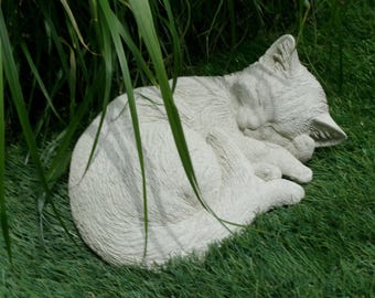 May include: A sleeping cat statue made of light gray stone. The cat is curled up on green grass, with its paws tucked in and head resting on its front paws. The statue is partially obscured by green blades of grass.