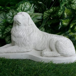 May include: A white, stone-like sculpture of a dog in a reclining position. The dog is lying on a rectangular base, with its head raised. The sculpture is set against a backdrop of green foliage and grass.