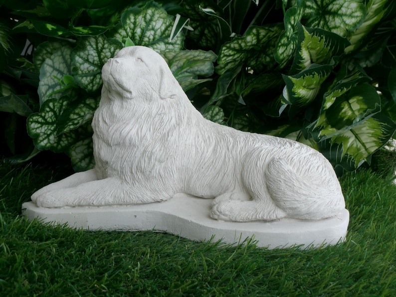 May include: A white, stone-like sculpture of a dog in a reclining position. The dog is lying on a rectangular base, with its head raised. The sculpture is set against a backdrop of green foliage and grass.