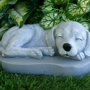 May include: A light gray sleeping puppy statue with closed eyes and a black nose. The dog is lying on a gray, rounded rectangular base. Green foliage is in the background.