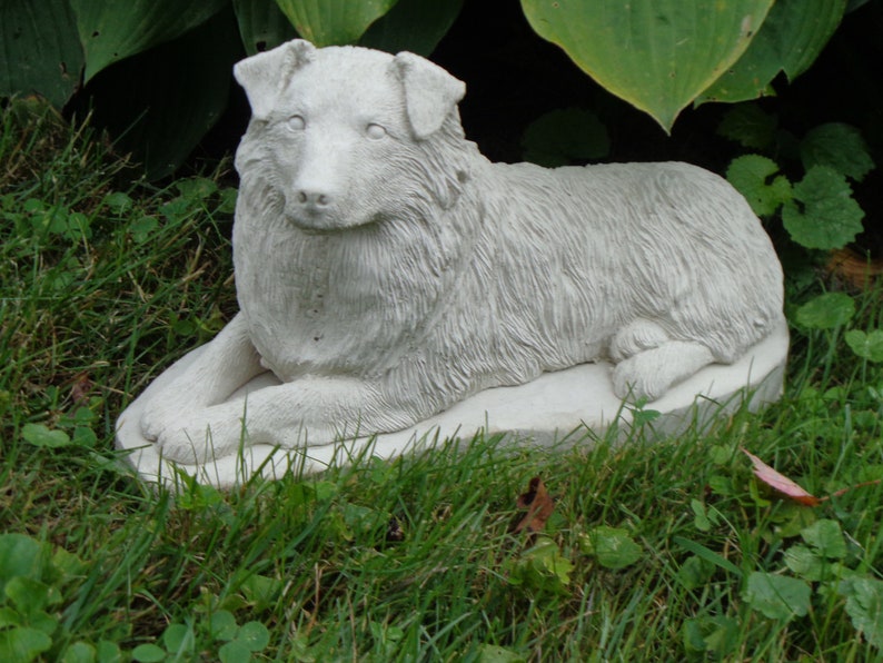 May include: A white concrete statue of a dog lying down. The dog is sculpted in a realistic style with fur details. The statue is placed on a grassy surface.
