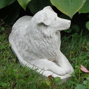 May include: White concrete statue of a dog sitting on a base. The dog is a long-haired breed with a fluffy tail and a long snout. The statue is in a grassy area with green leaves in the background.