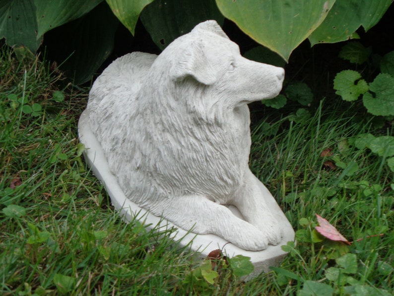 May include: White concrete statue of a dog sitting on a base. The dog is a long-haired breed with a fluffy tail and a long snout. The statue is in a grassy area with green leaves in the background.